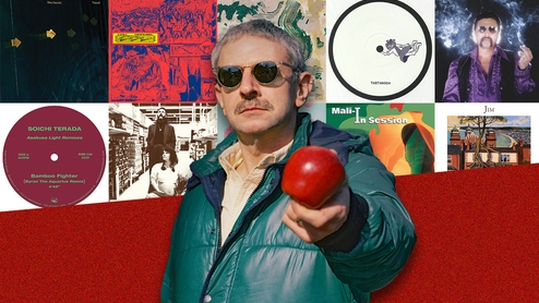 Ruf Dug in a green puffer jacket and sunglasses holding an apple in front of him, standing in front of a selection of artwork from his Selections