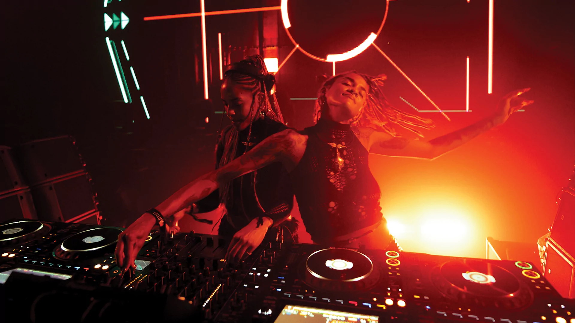 Photo of Coco & Breezy DJing behind the decks with orange and red lights